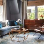 Sitting area with gray couch, brown leather chair, metal and stone coffee table, blue accent pillows, wood chest of drawers, window covered with taupe drapes