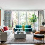 The living area featuring a couch upholstered in wide green and green-blue stripes, floor-to-ceiling windows that look out over trees, a pale taupe couch, a wood credenza and a large piece of art featuring rainbow-colored stripes
