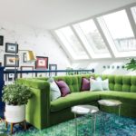 A sitting area featuring a green sectional couch, skylights, white walls, glass hexagonal tables and a shag rug in shades of green, blue and purple.