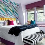 Guest room with a bed featuring a mustard colored headboard, white duvet and a black-and-white striped bench at the foot of the bed. The wallpaper behind the bed is a geometric pattern in shades of purple, blue, green and gray.