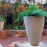 A natural-colored urn with a green plant in it. It's on a table and behind it are brick planters and green foilage.