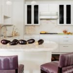 Looking into the kitchen we see white cabinets and an eat-in bar with bar stools covered in purple vinyl. There are eggplants on the white countertop.