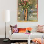 A white couch with throw pillows in oranges and purples. There is a large painting of tulips in a vase above the couch. The tulips are pink and red. There's also a cocktail table with a vase of orange flowers.