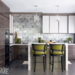 A view into a small kitchen with gray tile backsplash. There's a bar with two green barstools. Teo glass pendent lights hang above the bar.