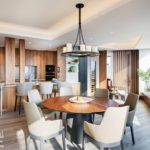 Round, walnut dining table surrounded by taupe colored chairs with a view into the walnut kitchen