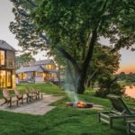 The back of the cottage featuring an expansive green lawn in front of a river. There are Adirondack chairs facing the river and surrounding a fire pit plus two chaise lounge chairs facing the river