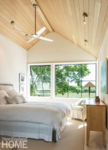 Master bedroom with a bed covered in white bedding. There's a ceiling fan hanging from the pitched roof over the bed. Windows look out on the green yard.