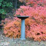 A gray bird bath with red fall foliage in the background.