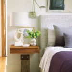 Close up shot of the side of a bed with a gray headboard and white and eggplant bedding. There's a floating nighstand with a lamp and a vase of hydrangeas.