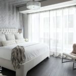 Master bedroom in white and light gray. There's a bed in white coverings, and a large window with white shears