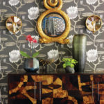 a buffet table in various woods sits in front of a wall with lotus-patterned wallpaper. There's a gold-framed mirror hanging on the wall.