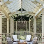 Cream-colored pergola houses four black chairs with white cushions that surround a wood table adored with a white vessel of white flowers