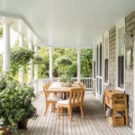 On the wraparound porch, you have a rectangular table made of light-colored wood and adorned with a plant. There's also a side table under a window.