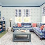 Living room in shades of blue and green. There's wallpaper on the walls, roman shades on the windows and a blue bookcase filled with books. A photo of waves hangs above the bookcase.
