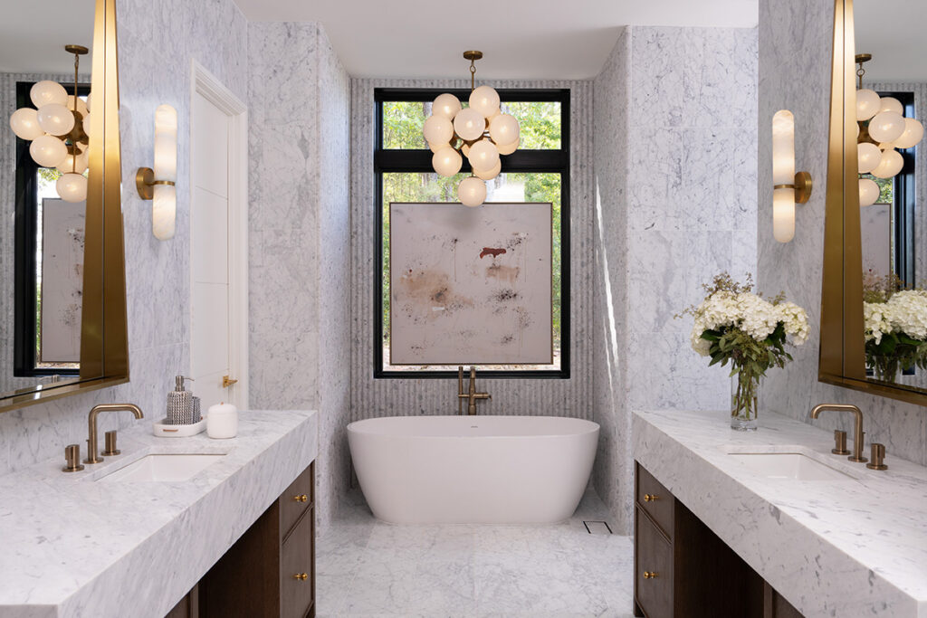 Luxurious modern bathroom with white marble walls and floors, featuring a freestanding white bathtub centered under a large window with black trim. Double vanities with marble countertops, brass fixtures, and dark wood cabinets add elegance to the space. Statement lighting includes a cluster chandelier and wall sconces, enhancing the bathroom's contemporary design.