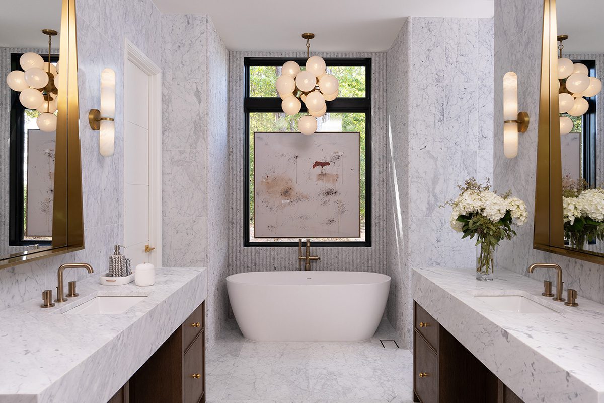 Luxurious modern bathroom with white marble walls and floors, featuring a freestanding white bathtub centered under a large window with black trim. Double vanities with marble countertops, brass fixtures, and dark wood cabinets add elegance to the space. Statement lighting includes a cluster chandelier and wall sconces, enhancing the bathroom's contemporary design.