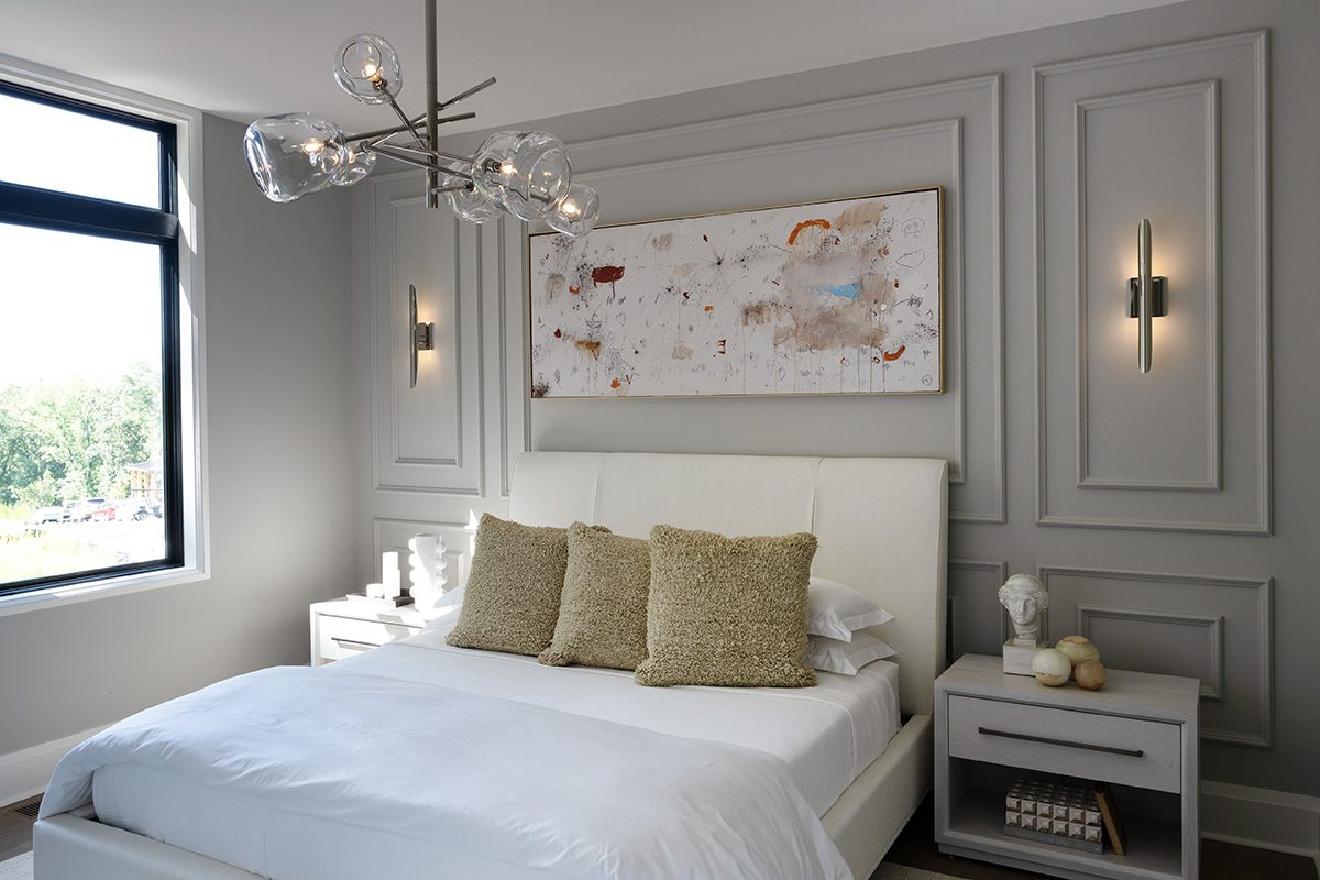 Modern bedroom with gray paneled walls, featuring a white upholstered bed with plush textured pillows. Above the bed hangs abstract artwork, flanked by sleek wall sconces for layered lighting. A contemporary chandelier with glass globes from Regina Andrew Lighting adds elegance and illumination to the room. Light-colored bedside tables hold decorative items, enhancing the room's serene, sophisticated ambiance.