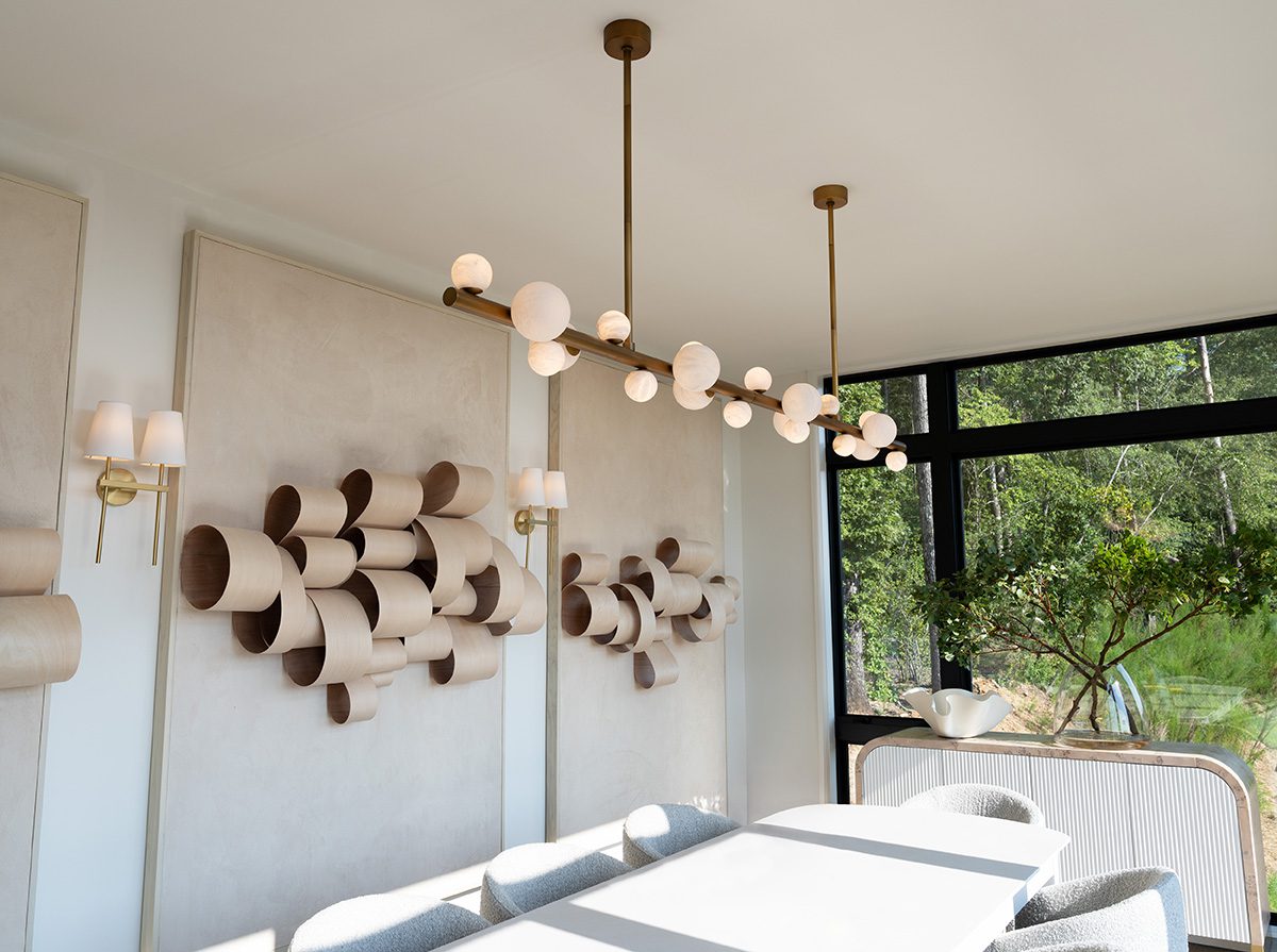 Contemporary dining room with minimalist decor, featuring a white table, upholstered chairs, sculptural wall art, brass chandelier with globe lights, and brass wall sconces, with natural light streaming in through large windows.