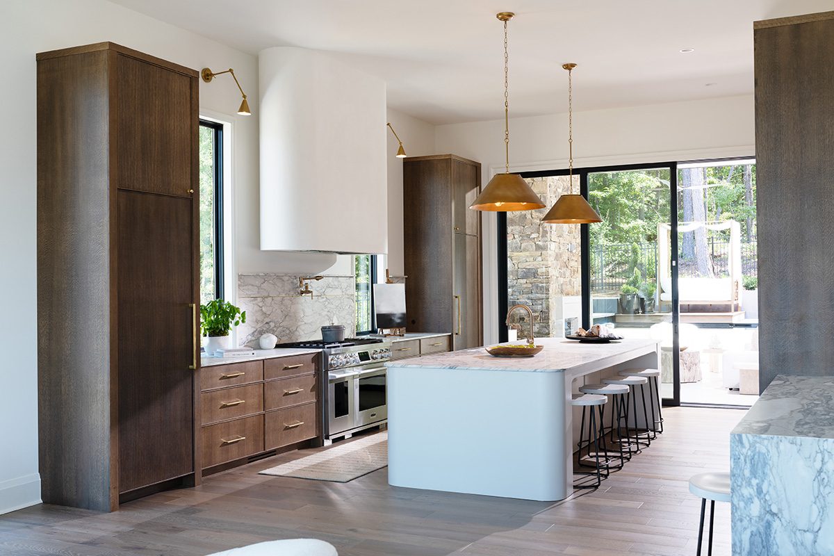 Modern kitchen with dark wood cabinets, white marble countertops, and a large central island featuring built-in seating and brass pendant lighting. The kitchen includes a professional stainless steel range with a white custom range hood and marble backsplash. Floor-to-ceiling windows open to an outdoor patio, providing natural light and a seamless indoor-outdoor flow, perfect for entertaining