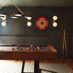 Stylish home billiard room with a modern pool table, dark walls, unique lighting fixtures, and vintage game decor, creating a sophisticated and cozy ambiance.