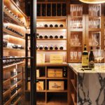 Close-up of a luxurious wine cellar featuring illuminated wooden shelves filled with wine bottles, glassware, and a marble tasting table with wine bottles and glasses