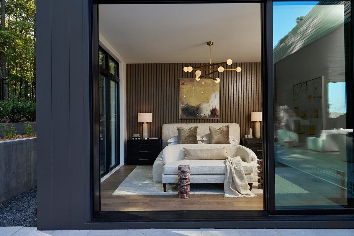 Modern bedroom with a cozy neutral palette featuring a plush bed, elegant beige sofa, textured wooden accent wall, abstract artwork, and a contemporary chandelier with globe lighting fixture by Regina Andrew. 