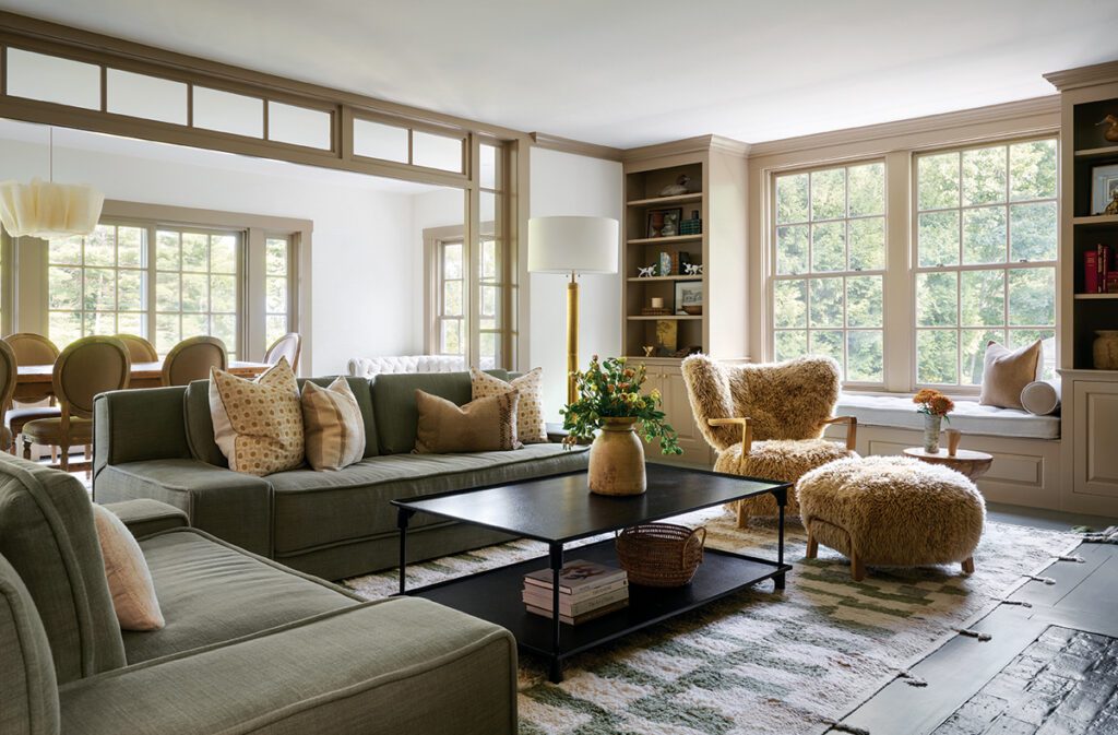 Bright and inviting living room featuring a sectional sofa with neutral throw pillows, a black metal coffee table, and cozy shearling chairs. The space includes built-in shelving, a window seat with cushions, and large windows providing natural light. A modern dining area is visible through a custom glass partition, creating an open yet defined layout.