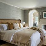 Elegant bedroom featuring a woven rattan bedframe with neutral bedding and layered throw pillows, a grasscloth wallpaper accent wall, and a cozy fireplace. The space includes an arched doorway leading to a seating area, subtle decor with natural textures, and a calming color palette for a serene retreat.