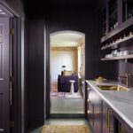 Moody and luxurious butler's pantry featuring rich plum-colored cabinetry, a marble countertop, and brass hardware. The space includes open shelving for display, an arched doorway leading to a cozy living area, and a vintage-inspired runner that adds warmth and character.