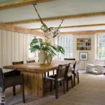 Rustic and elegant dining room featuring a reclaimed wood table paired with leather chairs and a sculptural chandelier. The space includes exposed wooden beams, large grid windows providing natural light, and built-in shelves displaying decor. A natural fiber rug and neutral tones create a warm and inviting ambiance.