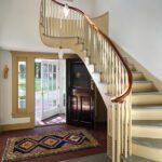 Elegant entryway featuring a sweeping curved staircase with a polished wooden handrail, a classic black front door, and French doors leading to a lush outdoor view. The space is accented by a vibrant patterned rug, brick flooring, and soft natural light, blending traditional charm with timeless design.