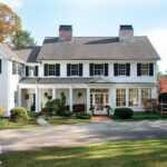 Stunning colonial-style home with classic white siding, black shutters, and brick chimneys, surrounded by manicured landscaping and a lush green lawn. The inviting exterior features a charming covered porch, large windows, and a scenic backdrop of mature trees and vibrant foliage, blending timeless elegance with picturesque New England charm.
