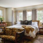 Charming guest bedroom featuring a neutral upholstered bed with layered pillows and a cozy throw, large windows with flowing drapery, and warm natural light. The room is accented by a vintage floral-patterned loveseat, wooden nightstands, and a writing desk with fresh greenery, creating a serene and inviting retreat.