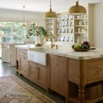 Bright farmhouse-style kitchen featuring a large wooden island with a marble countertop, a farmhouse sink with brass fixtures, and open shelving displaying white dishware. The space is illuminated by brass pendant lighting and natural light from large glass doors leading to an outdoor view. A vintage- rug adds warmth and texture to the elegant design.