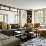 Bright and inviting living room featuring a sectional sofa with neutral throw pillows, a black metal coffee table, and cozy shearling chairs. The space includes built-in shelving, a window seat with cushions, and large windows providing natural light. A modern dining area is visible through a custom glass partition, creating an open yet defined layout.