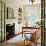 Elegant home office featuring built-in shelving, a fireplace with a painted mantel, and a large window flooding the space with natural light. The room includes a wooden desk with a sculptural table lamp, a vintage-style armchair, and a woven rocker with a cozy throw. Soft neutral tones and curated decor create a serene and functional workspace.