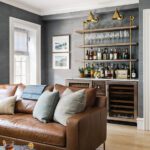 Chic home bar setup featuring open wooden shelves with brass accents, stocked with an assortment of spirits and glassware. The bar area includes built-in cabinetry, wine storage, and artwork by Sarah Madeira Day, creating a stylish and functional space. A cozy leather sofa with patterned throw pillows enhances the inviting atmosphere.