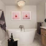 Elegant primary bathroom featuring a freestanding soaking tub with sleek chrome fixtures, accented by modern pink artwork and a sparkling crystal chandelier. The space includes marble flooring, a wooden vanity with a quartz countertop, and thoughtful decor, creating a luxurious and tranquil retreat.
