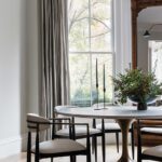 Bright and sophisticated dining area located in a bay window on the parlor level, featuring a round table with a sculptural metallic base, modern upholstered chairs, and minimalist black candleholders. The space is accented by neutral drapery, an antique mirror, and natural light streaming through the large window, blending formal elegance with family-friendly design." Bright and sophisticated dining area located in a bay window on the parlor level, featuring a round table with a sculptural metallic base, modern upholstered chairs, and minimalist black candleholders. The space is accented by neutral drapery, an antique mirror, and natural light streaming through the large window, blending formal elegance with family-friendly design."