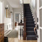 Bright and elegant Boston home entryway featuring a staircase with a dark patterned carpet runner, light hardwood floors, white paneled walls, and gold-accented wall sconces. A hallway leads to a cozy living space with a hint of warm-toned decor.