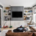 Cozy and functional family room featuring a built-in media wall with white cabinetry, open shelving for books and decor, and a mounted flat-screen TV. The space includes a comfortable leather sectional, a modern desk nook with a chair for a compact workspace, and woven storage baskets for toys. Dark gray textured walls and natural light from large windows create a warm and inviting atmosphere.