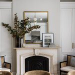 Stylish living area featuring a classic marble fireplace with a black cast-iron insert, flanked by built-in white cabinets with brass hardware. The mantel is adorned with a gold-framed mirror, a vase with seasonal branches, and framed art. Cane armchairs with plush cushions and light-toned walls create a warm and inviting atmosphere.