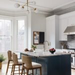 Bright and inviting kitchen featuring a navy island with a marble countertop, paired with woven barstools for a stylish yet functional seating area. The space boasts a bay window that floods the room with natural light, a modern brass chandelier with a decorative ceiling medallion, and high-end stainless steel appliances. Designed with flexibility in mind, the open layout leaves room for future family needs.
