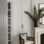 Elegant home design featuring hidden storage behind a sleek panel near the kitchen and living area. The space includes built-in shelving for barware and toys, a white cabinet with brass hardware, a decorative mantel with a stylish vase, and a cozy cane chair paired with stacked books, blending functionality and timeless style.