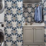 Charming laundry room featuring blue and white pineapple-patterned wallpaper, a compact sink with light gray cabinetry, and open shelving with woven storage baskets. The space includes a stacked washer and dryer and a hanging rod for clothing, combining functionality with playful design elements.