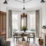 Elegant living and dining space in a Boston brownstone featuring a modern three-armed AERIN light fixture hanging from a decorative plaster ceiling medallion. The room includes a round dining table with sleek chairs, a large antique mirror, tall windows with neutral drapery, and leather-accented stools, blending historic charm with contemporary design.