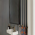 Bold and stylish powder room featuring navy and white vertical striped wallpaper, a sleek rectangular mirror, and a compact white pedestal sink with antique brass fixtures. A colorful floral towel adds a pop of contrast, enhancing the room’s vibrant and sophisticated design.