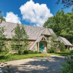 Exterior view of a charming Cotswalds-style cottage with a pitched roof, stone facade, lush landscaping, and a circular gravel driveway under a bright blue sky with scattered clouds.