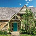 Front entrance of a Cotswalds-style cottage featuring a vibrant teal door, stone facade, peaked gable, and lush greenery under a bright blue sky with scattered clouds.