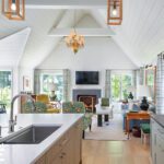 Bright and airy open-concept living room and kitchen with vaulted shiplap ceilings, an elegant chandelier, modern pendant lighting, a large island with a sink, and a cozy fireplace surrounded by comfortable seating.